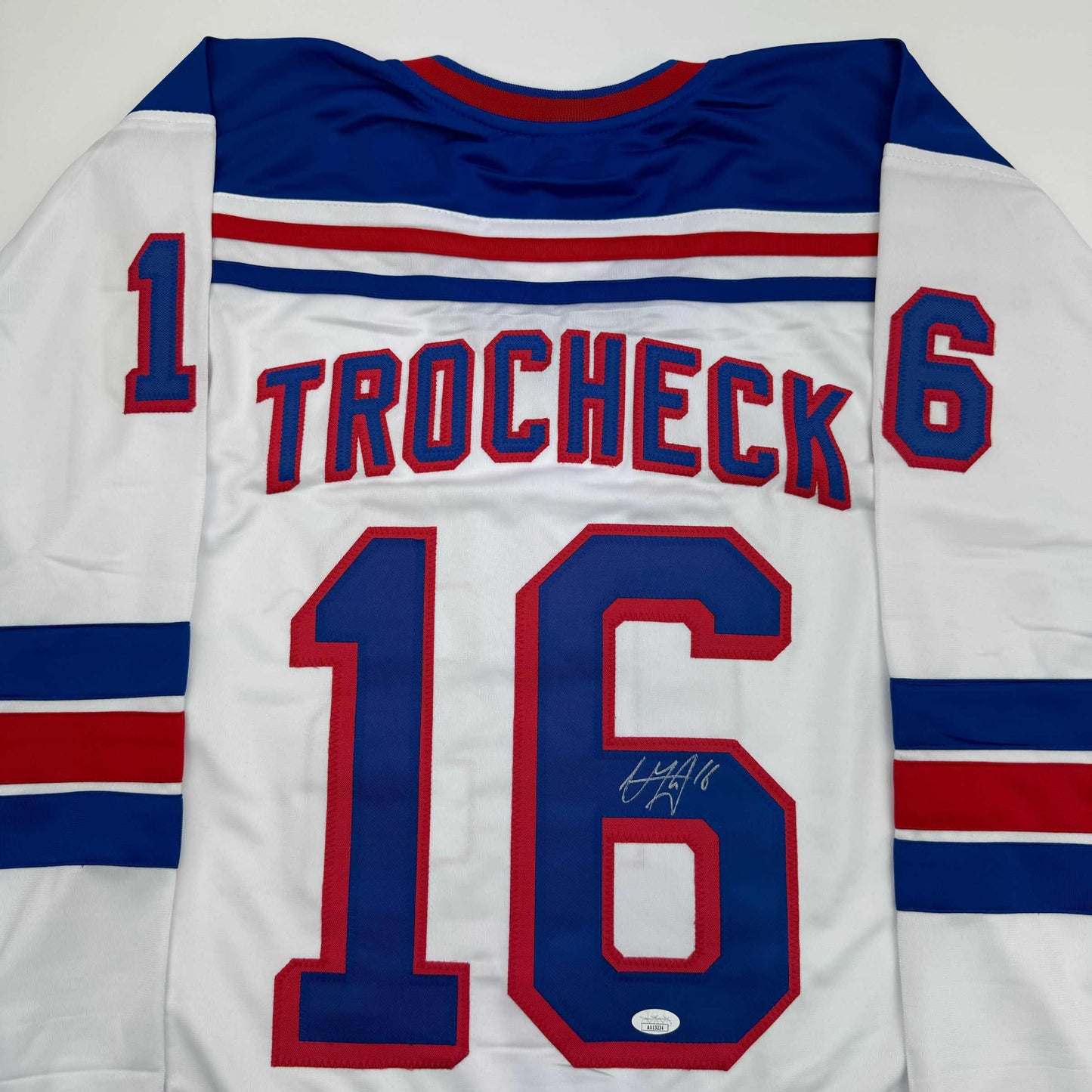 White hockey jersey with navy and red striping, displaying "TROCHECK" and number 16 on back, with visible autograph.