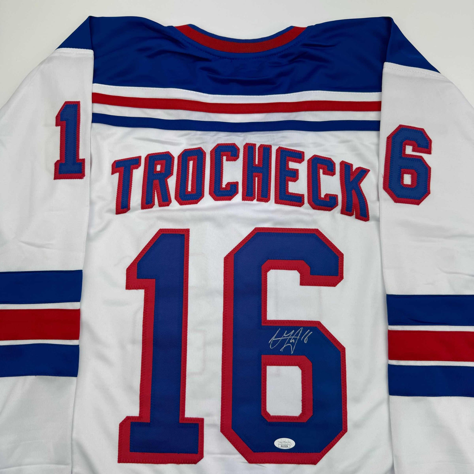 White hockey jersey with navy and red striping, displaying "TROCHECK" and number 16 on back, with visible autograph.