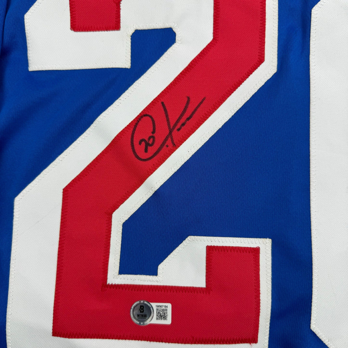 Close-up of blue New York Rangers hockey jersey number 16 with red stripe and handwritten signature, showing authentication hologram at bottom.