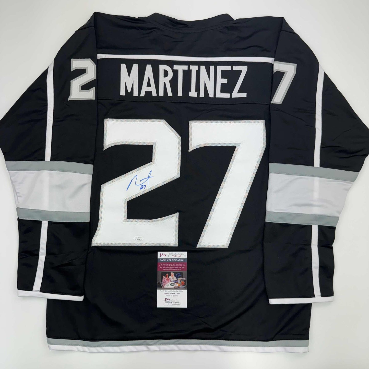 Back view of black LA Kings hockey jersey number 27 with white "MARTINEZ" nameplate and signature, displayed flat with JSA authentication certificate visible at bottom.