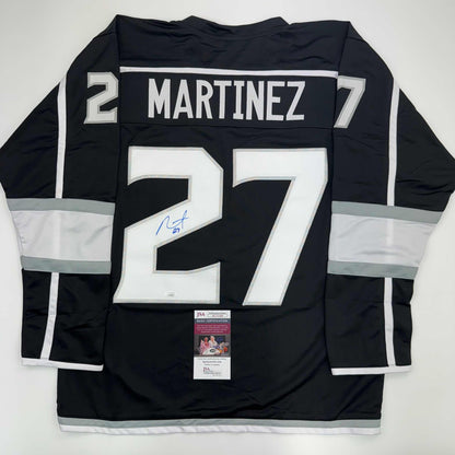 Back view of black LA Kings hockey jersey number 27 with white "MARTINEZ" nameplate and signature, displayed flat with JSA authentication certificate visible at bottom.