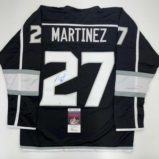 Back view of black LA Kings hockey jersey number 27 with white "MARTINEZ" nameplate and signature, displayed flat with JSA authentication certificate visible at bottom.