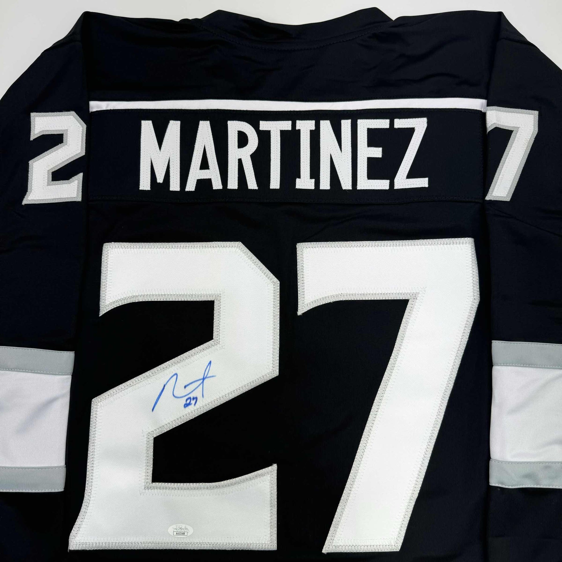 Back view of black hockey jersey displaying "MARTINEZ 27" with white stitched numbers and a blue autograph on the number seven.
