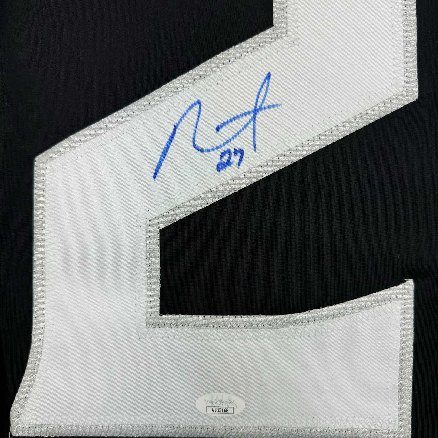 Close-up of white jersey sleeve showing blue handwritten signature "1/27" and authentication label at bottom.