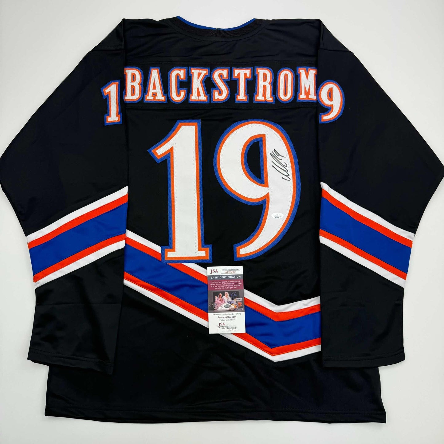 Black hockey jersey with blue and red striping displaying "BACKSTROM 19" and JSA authentication certificate attached to front.