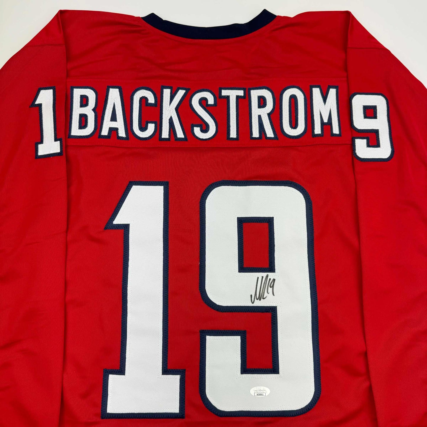 Back view of red hockey jersey displaying "BACKSTROM 19" in white and navy lettering with autographed number 19.
