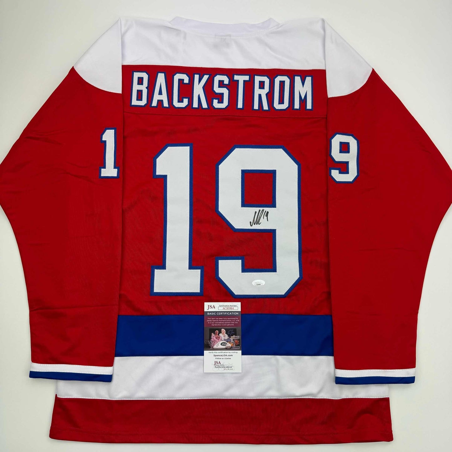 Red hockey jersey with white and blue trim displays "BACKSTROM" and number 19 with signature and JSA authentication certificate attached.