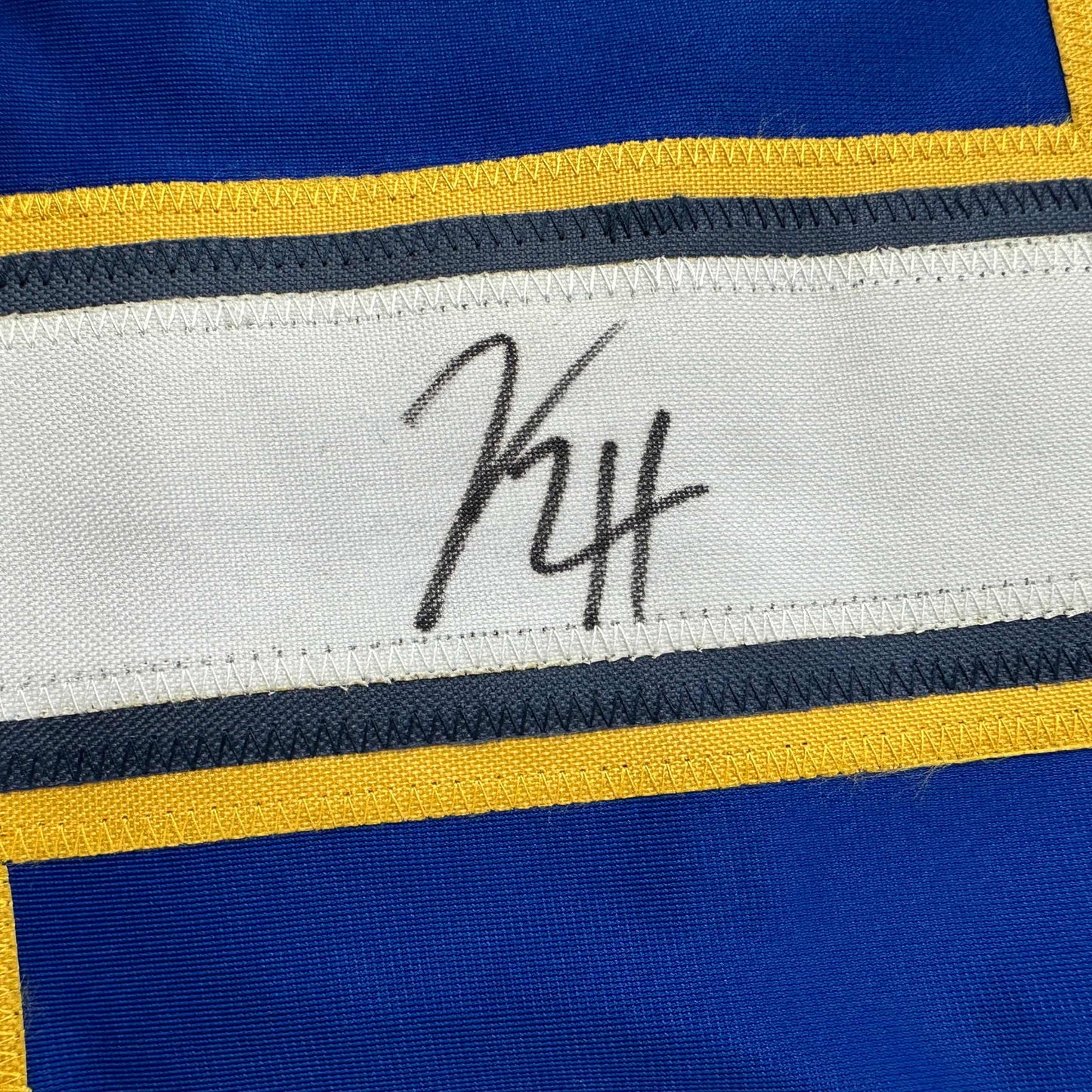 Close-up of autographed white stripe on blue and yellow hockey jersey showing handwritten signature "KH".