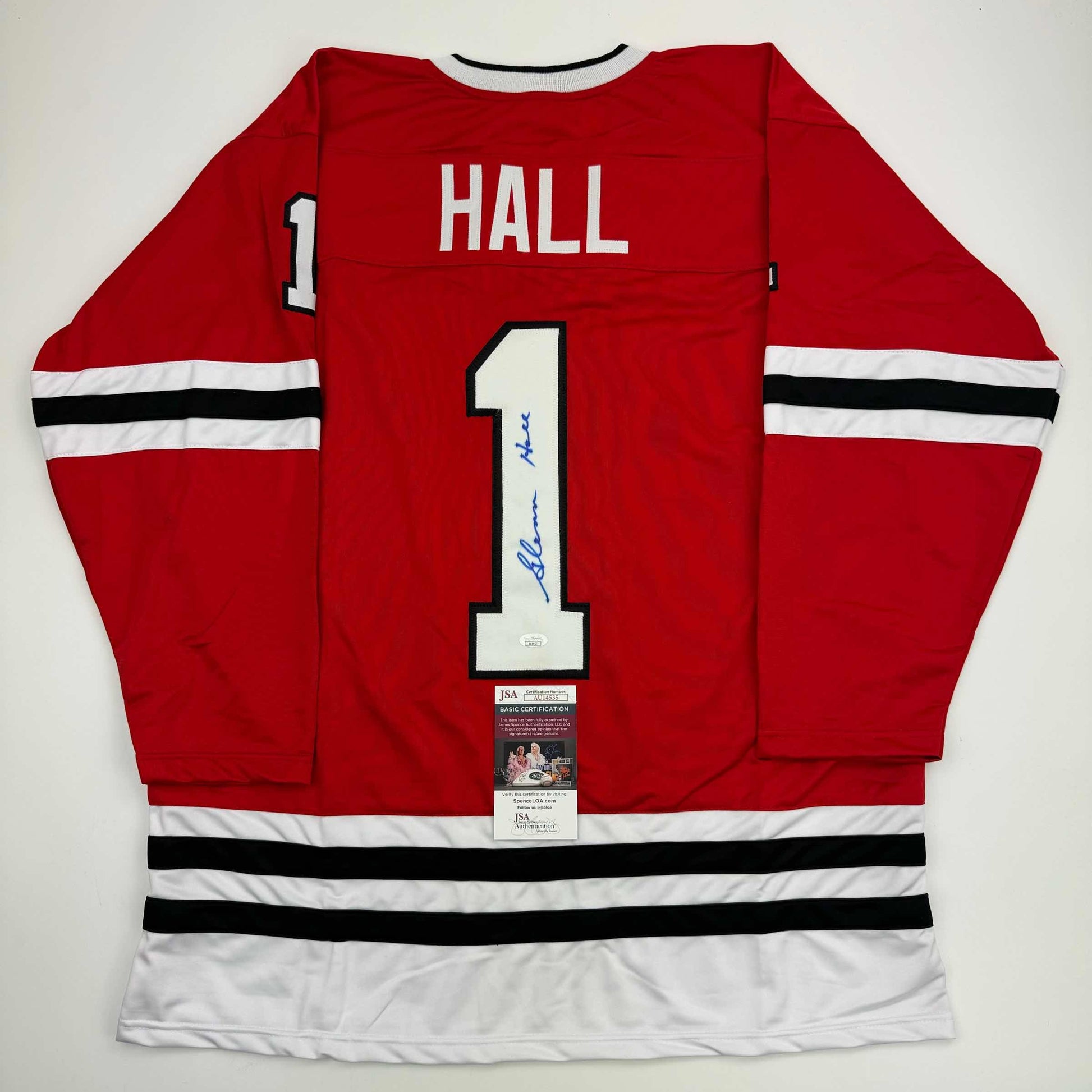 Back view of red Chicago Blackhawks jersey number 1 with "HALL" nameplate, white stripes, and blue autograph, displayed flat with JSA certification labels.