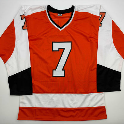 Flat lay of red, white, and black hockey jersey with large white number 7 on chest, displayed against white background.