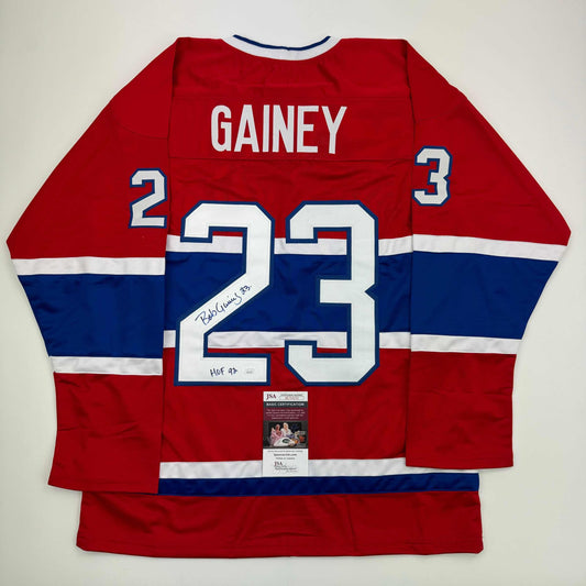 Red, white, and blue Montreal Canadiens hockey jersey with "GAINEY" and number 23 displayed flat, showing back view with JSA authentication certificate visible.