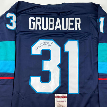 Back view of navy blue hockey jersey with "GRUBAUER" and number 31 in white and blue lettering, signed in black ink, with JSA authentication label visible at bottom.