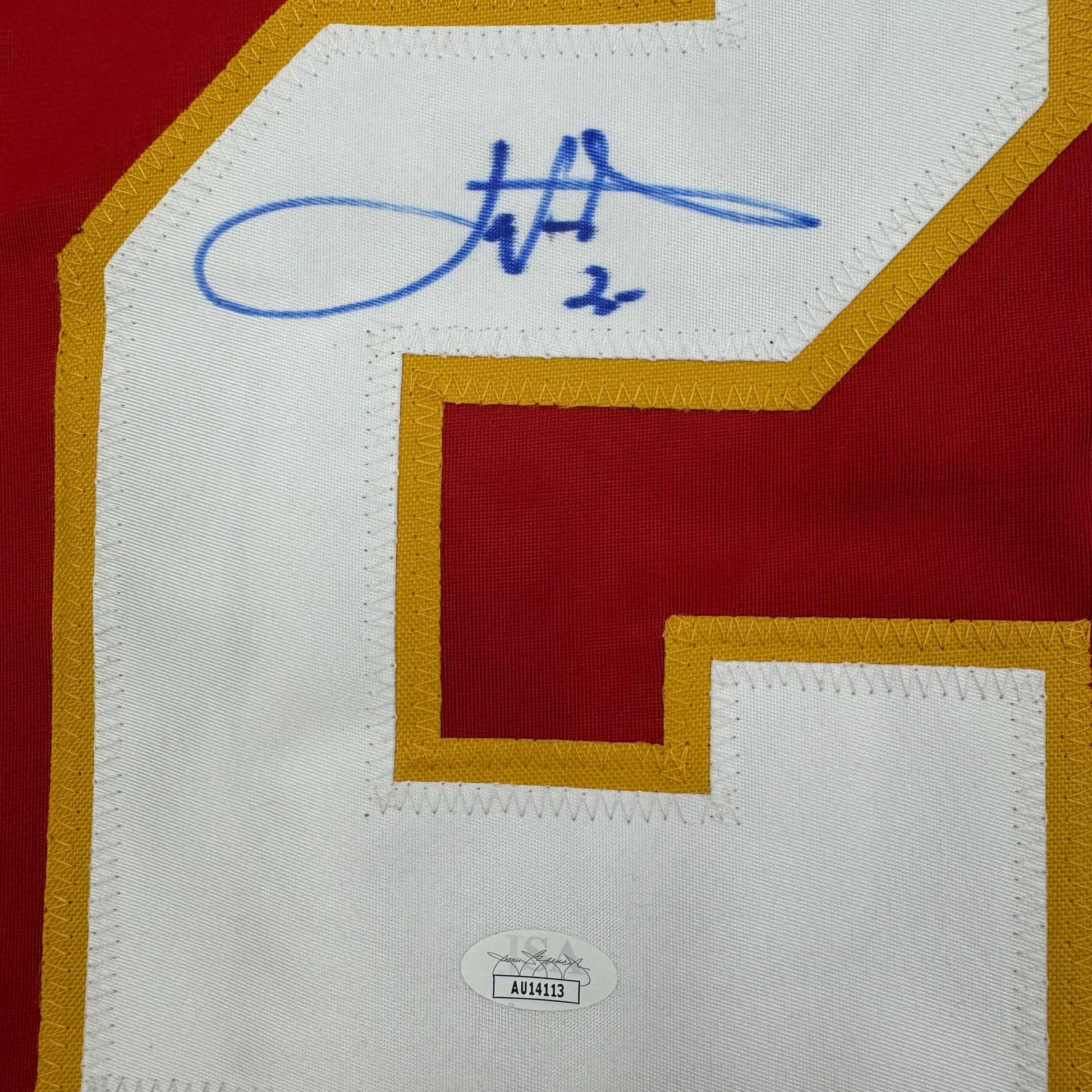 Close-up of autographed white jersey panel with blue signature and "2x" notation, gold and red borders visible, JSA authentication sticker at bottom.