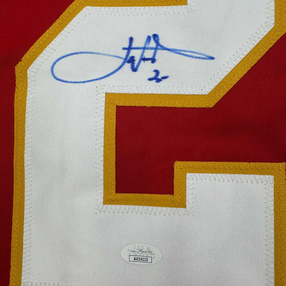 Close-up of autographed white jersey panel with blue signature and "2x" notation, gold and red borders visible, JSA authentication sticker at bottom.