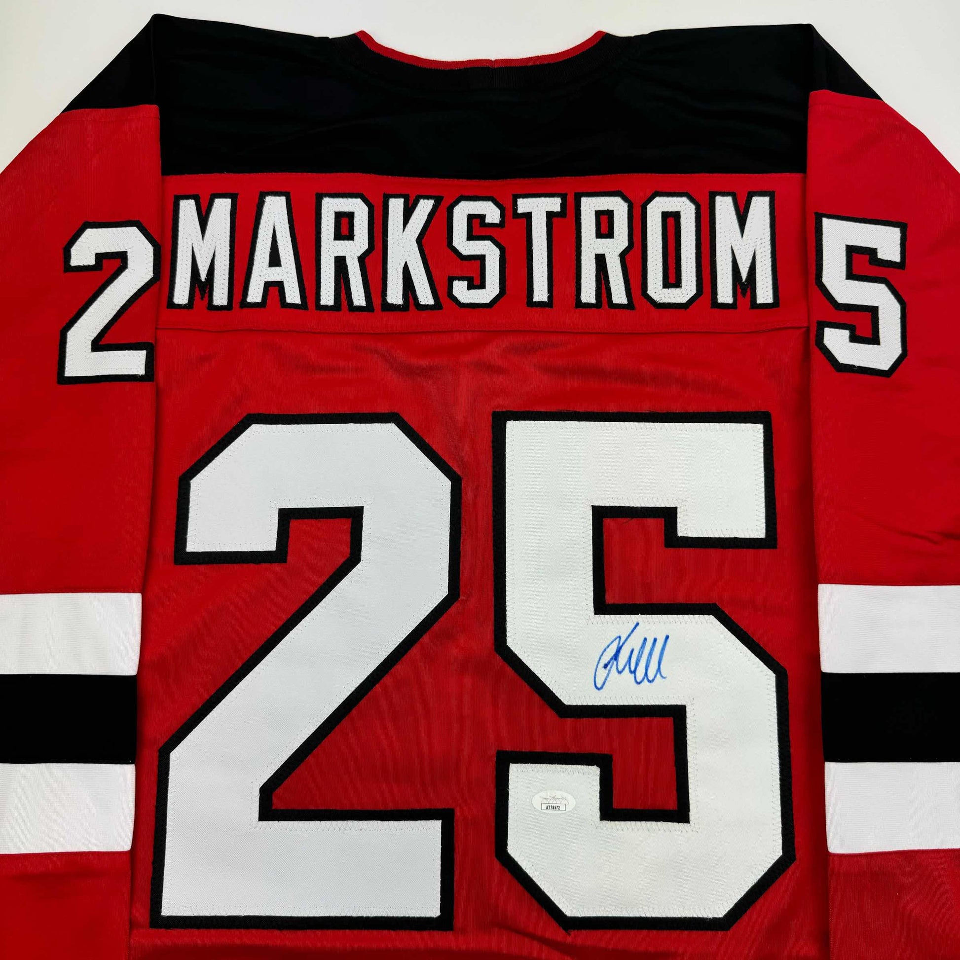 Back view of red hockey jersey with white numbers 25, "MARKSTROM" nameplate, black and white horizontal stripes, and blue autograph on number.