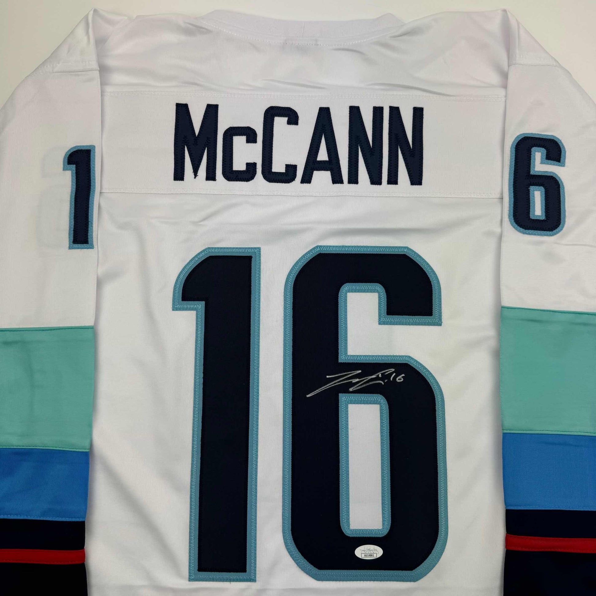 Back view of white hockey jersey displaying "McCANN" and number 16 with signature, colored stripe trim.