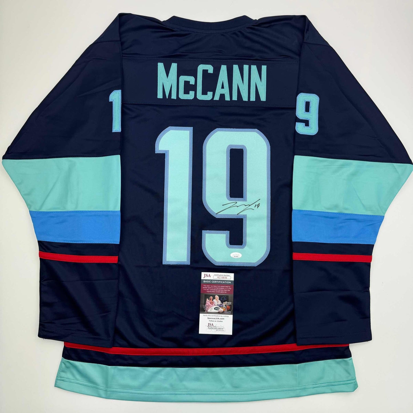 Reverse view of navy hockey jersey number 19 with "McCann" nameplate, light blue and royal blue horizontal stripes, signed with JSA authentication certificate visible.