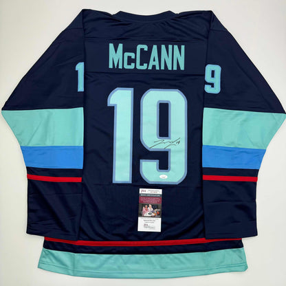 Reverse view of navy hockey jersey number 19 with "McCann" nameplate, light blue and royal blue horizontal stripes, signed with JSA authentication certificate visible.