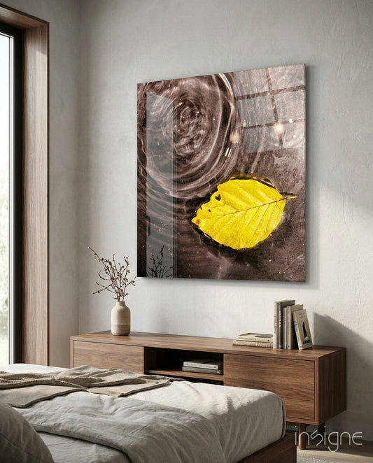 Glass wall art depicting a bright yellow leaf floating on dark rippled water, hung above a wooden bedroom console.