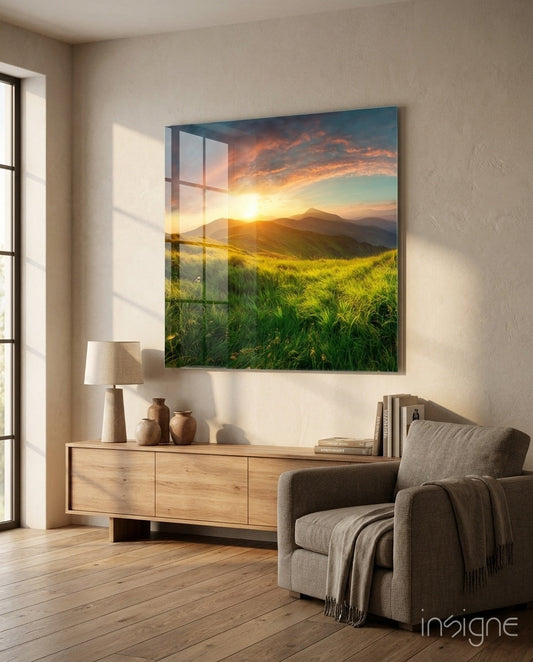Glass wall art depicting a sunset over green grassland with mountains, displayed above a wooden dresser in a modern living room.