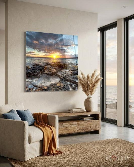 Square glass wall art depicting rocky coastline at golden sunset, displayed above wooden console table in coastal interior.