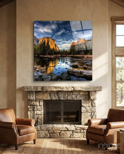 Square glass wall art featuring mountain valley with river, golden cliffs, and evergreen trees displayed above fireplace.