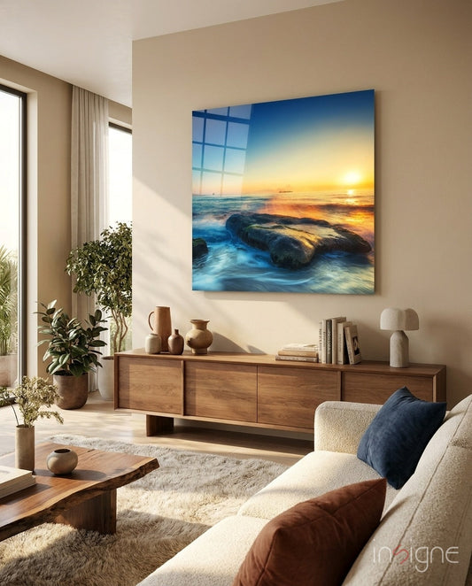 Rocky Shore Ocean Sunset Glass Wall Art displayed above a wooden credenza in a beige living room with coastal décor.