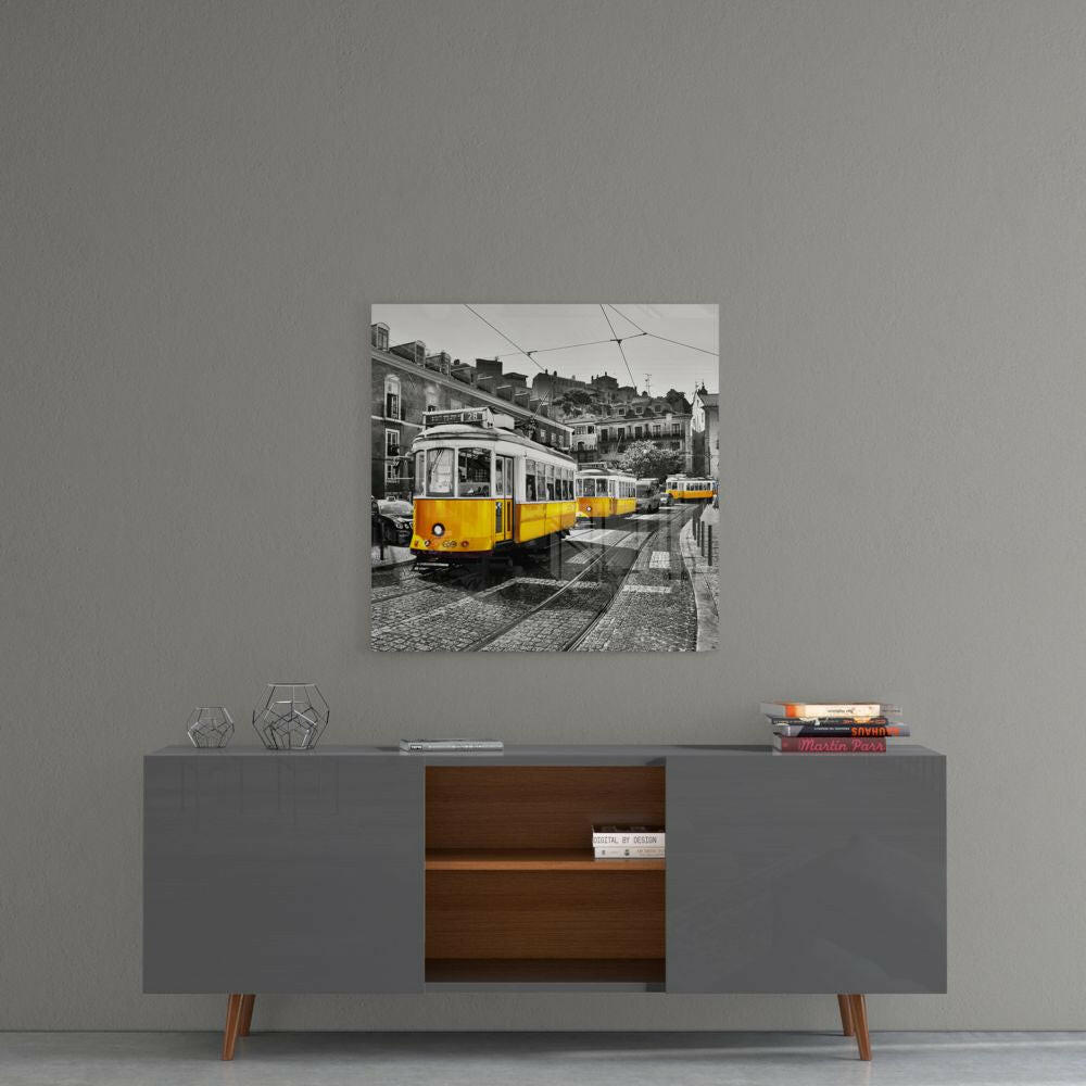 Square glass wall art mounted on gray wall above sideboard, showing yellow and white tram on cobblestone Lisbon street with historic buildings.