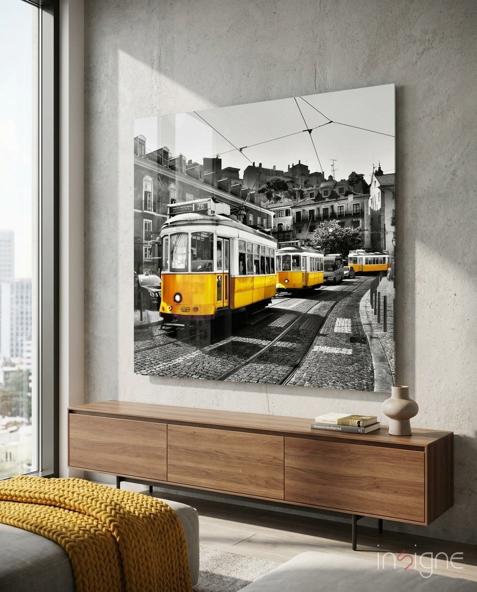 Yellow trams on cobblestone street mounted as glass wall art above wooden console in modern living room with concrete wall.