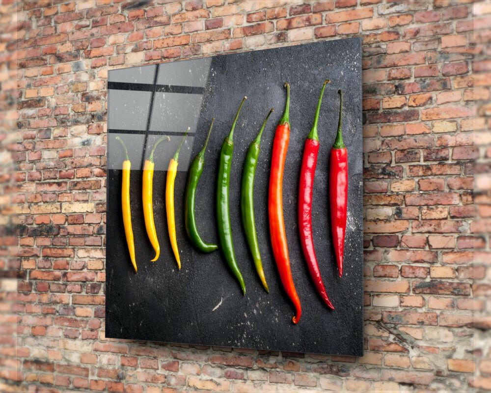 Glass wall art panel mounted on brick displaying nine chili peppers in yellow, green, and red arranged in color-gradient rows against dark background.
