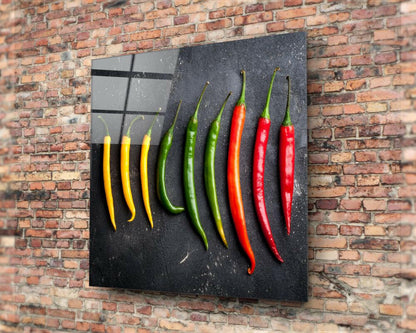 Glass wall art panel mounted on brick displaying nine chili peppers in yellow, green, and red arranged in color-gradient rows against dark background.