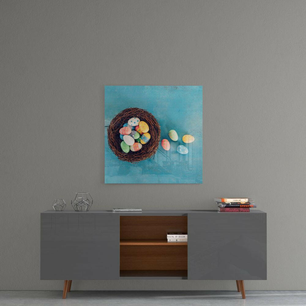 Square glass wall art depicting a dark woven nest filled with colorful Easter eggs on turquoise background, displayed above modern sideboard.