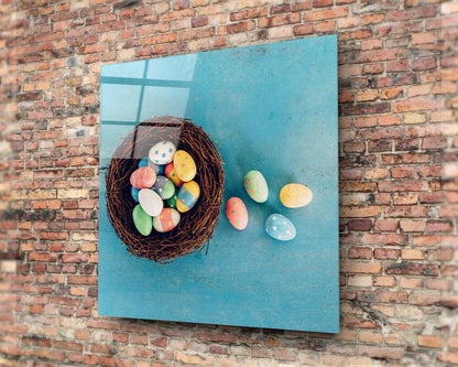 Glass wall art mounted on red brick showing a woven nest with colorful painted eggs on turquoise background, with eggs scattered beside it.