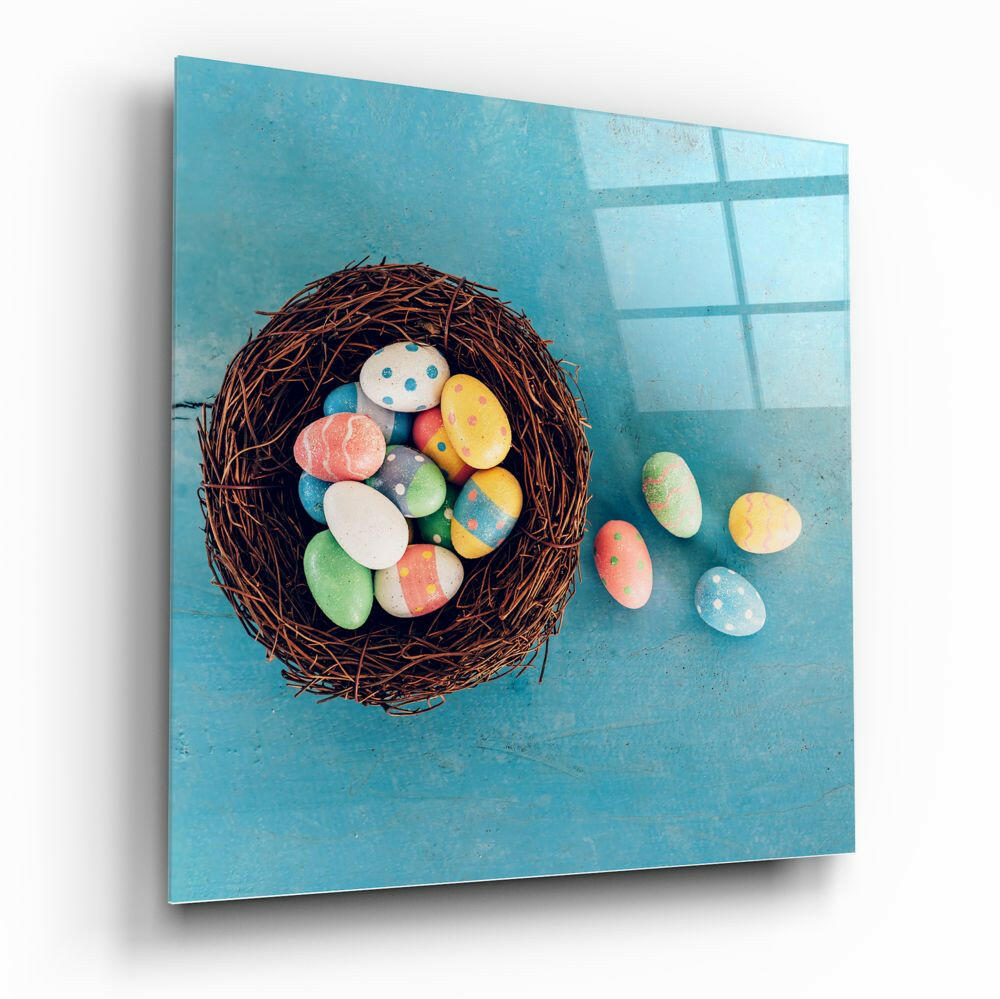 Colorful painted Easter eggs spilling from a brown twig nest onto turquoise painted wood background with window panes, displayed on glass wall art at an angle.