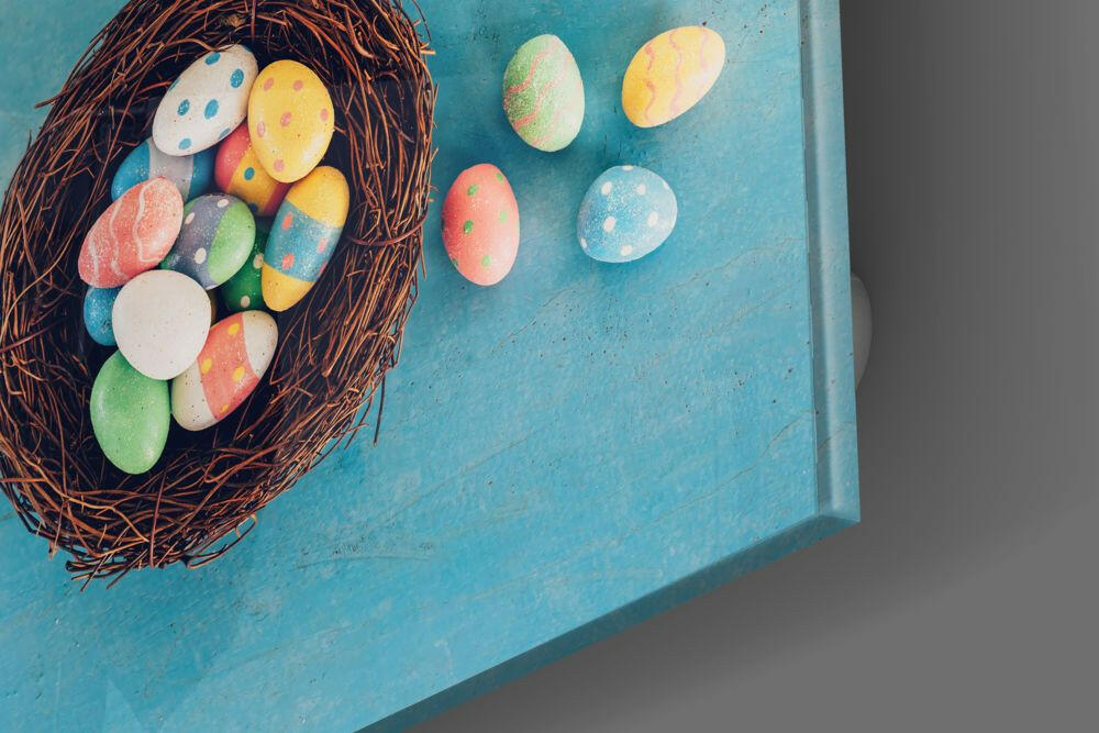 Overhead view of colorful hand-painted Easter eggs in a brown woven nest on turquoise background, with additional eggs scattered beside it.