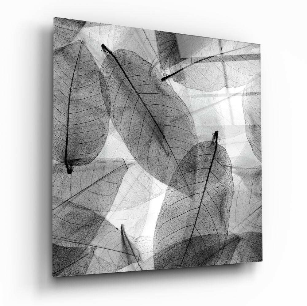 Angled view of glass wall art displaying overlapping skeletal leaf silhouettes in black and white, mounted on white background.