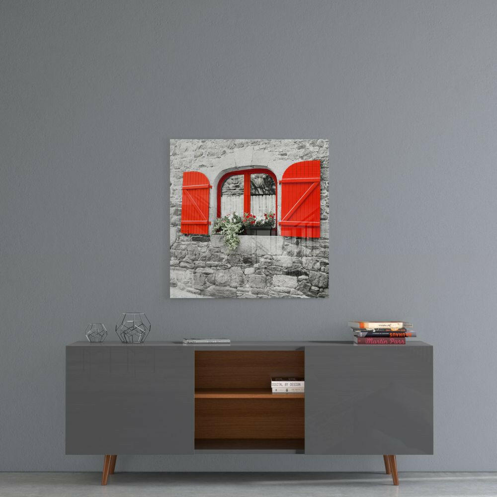 Square glass art print depicting a stone building facade with bright red shutters and window frame, displayed above a modern console table.