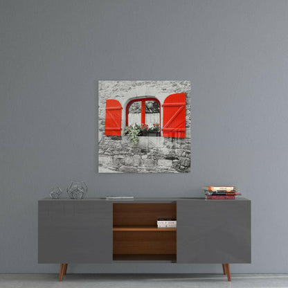 Square glass art print depicting a stone building facade with bright red shutters and window frame, displayed above a modern console table.