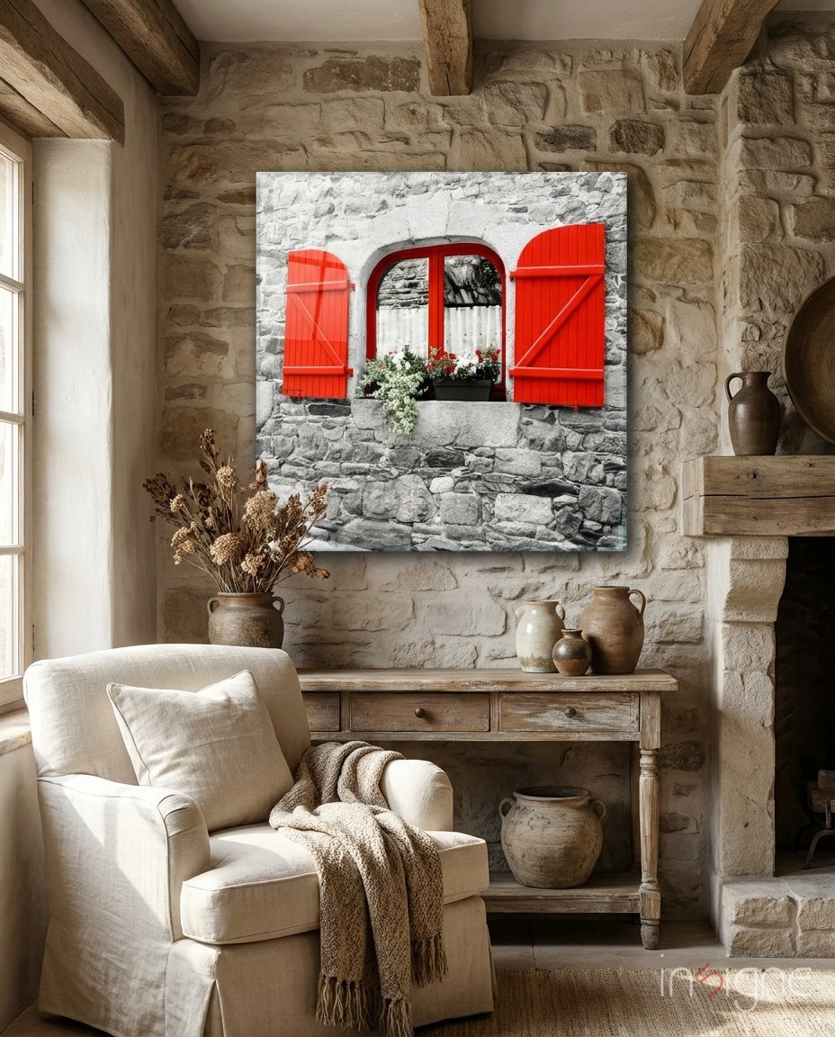 Red shutters and window with flowering box on stone wall displayed above wooden console table in rustic interior.