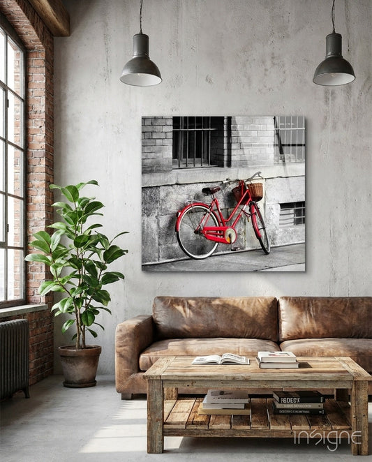 Red bicycle with basket leaned against gray brick building wall, displayed as canvas art above brown leather sofa in industrial loft interior.
