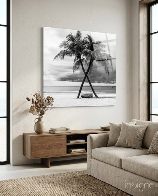 Black and white glass wall art of two crossed palm tree trunks on beach, displayed above wooden console in minimalist living room.
