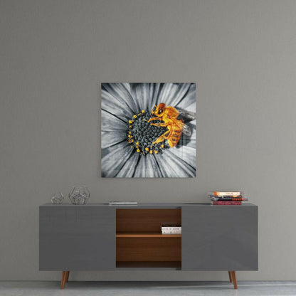 Square glass wall art depicting a bee on a flower with selective color technique, white petals and yellow center details, mounted on gray wall above wooden sideboard.