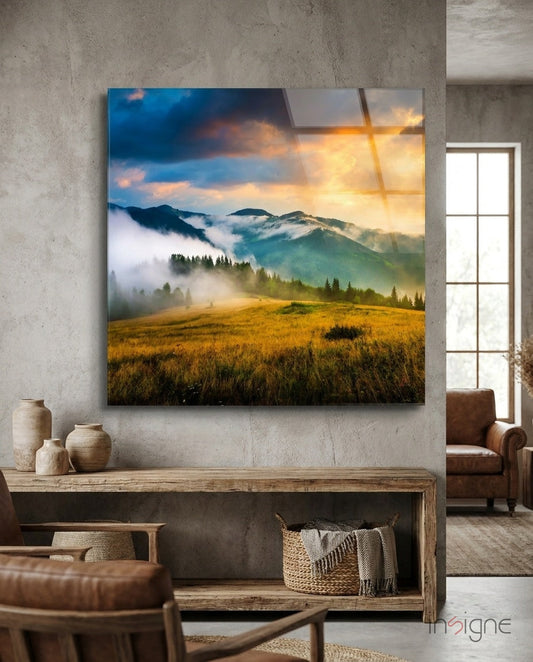 Glass wall art depicting misty mountains, golden meadow, and sunset sky displayed above wooden console in contemporary living room.