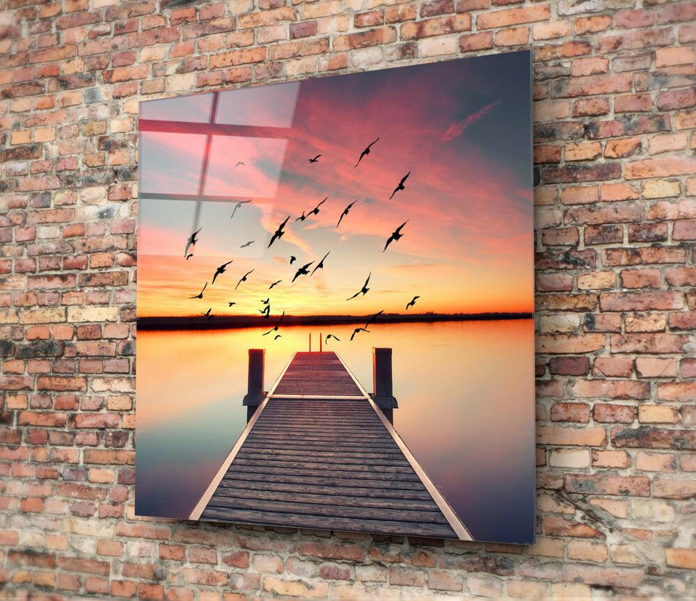 Glass wall art mounted on brick showing wooden dock extending into sunset water with flying birds silhouetted against orange sky.