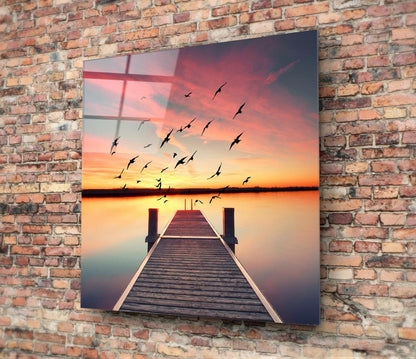 Glass wall art mounted on brick showing wooden dock extending into sunset water with flying birds silhouetted against orange sky.