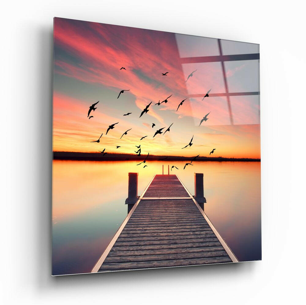 A wooden dock extends into a calm lake beneath a vibrant sunset sky with silhouetted birds in flight, displayed on glass wall art.