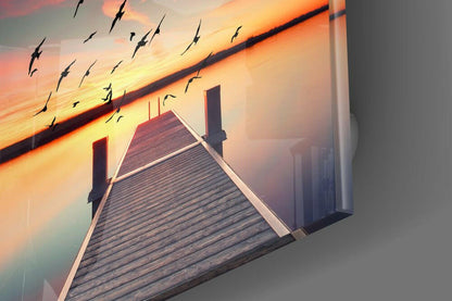 Wooden dock extending into sunset sky with flying birds and distant cityscape silhouette on glass wall art.