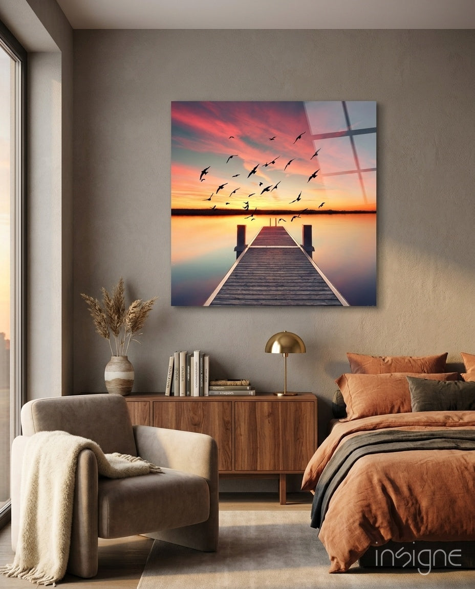 Red Dock glass wall art displaying a wooden pier at sunset with flying birds and pink-orange sky, hung in a bedroom above a wooden dresser.