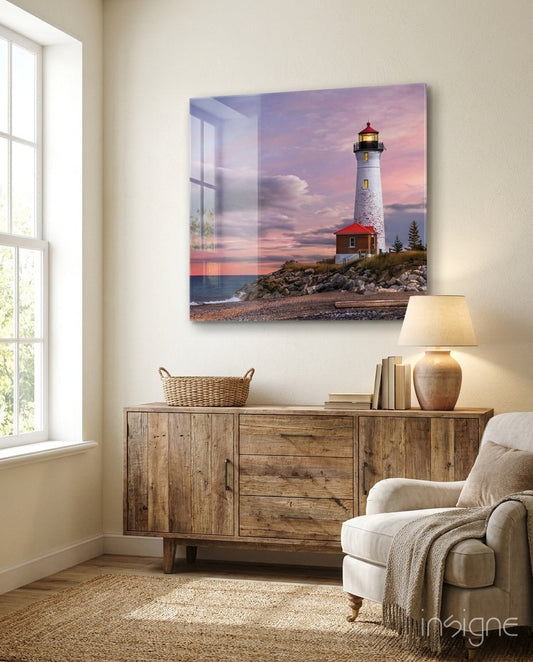 Glass wall art depicting a white lighthouse with red base on rocky shore at pink sunset, displayed above wooden dresser in beige living room.