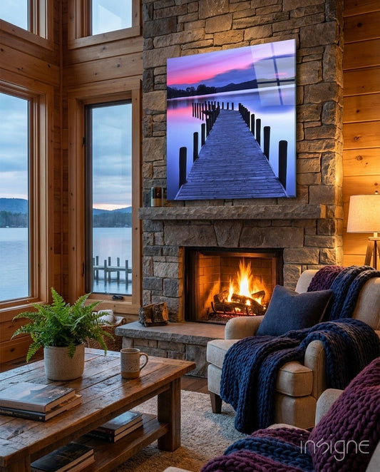 Glass wall art displaying a wooden jetty at sunset with pink and purple sky, hung above fireplace in lakeside cabin interior.